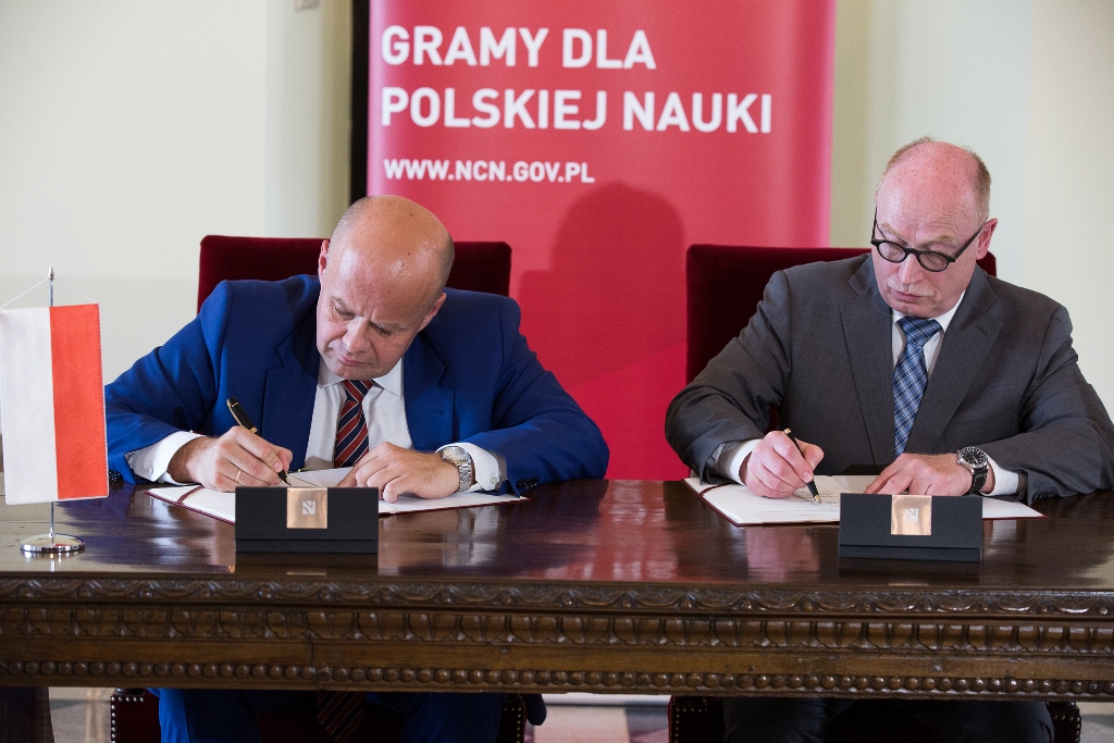 Zbigniew Błocki, dyrektor NCN i Martin Stratmann, prezydent MPG podpisują porozumienie w sprawie utworzenia w Polsce sieci Centrów Doskonałości Naukowej Dioscuri. Panowie siedzą za zabytkowym stołem, na którym stoi proporczyk z polską flagą. Za plecami siedzących - czerwony rollup Narodowego Centrum Nauki z hasłem: Gramy dla polskiej nauki. Autor zdjęcia: Michał Niewdana