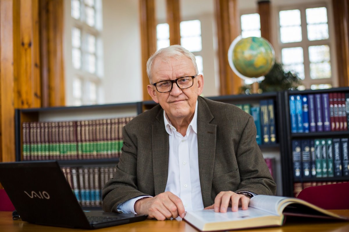 Prof. Juliusz Gardawski - zdjęcie portretowe na tle półki z książkami. Na stoliku przed profesorem leży laptop i otwarta książka.