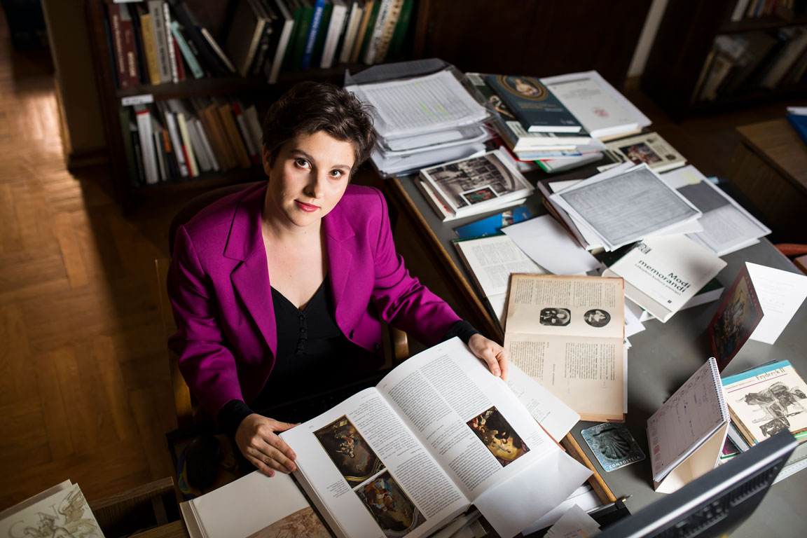 dr Emilia Kłoda siedzi przy biurku, na którym leży dużo książek i dokumentów. Przed sobą trzyma otwartą książkę z reprodukcjami obrazów Johanna Christopha Lischki.