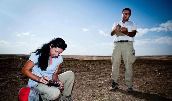 Agata Smilgin klęczy na ziemi wprowadzając do przenośnego urządzenia dane z GPS, obok stoi Rafał Koliński z notatnikiem w rękach.