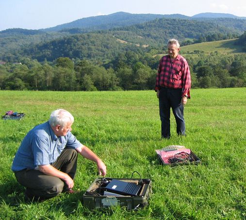 Na górskiej łące znajduje się dwóch mężczyzn. Pierwszy siedzi na trawie obok rozłożonego sprzętu badawczego. Drugi stoi niedaleko.