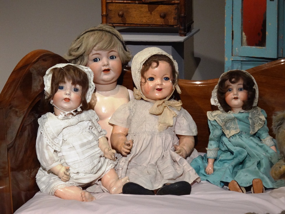 Cztery lalki w jasnych sukienkach, posadzone na łóżku. Lalki pochodzą z kolekcji Marka Sosenko. Fotografia autorstwa Doroty Żołądź-Strzelczyk.