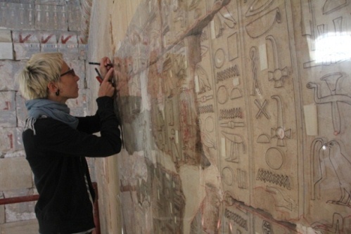 O zdobioną ścianę świątyni oparta jest przezroczysta tafla z pleksi. Anastasiia Stupko-Lubczyńska stoi przed ścianą i za pomocą markera przerysowuje na pleksi zdobienia ze ściany