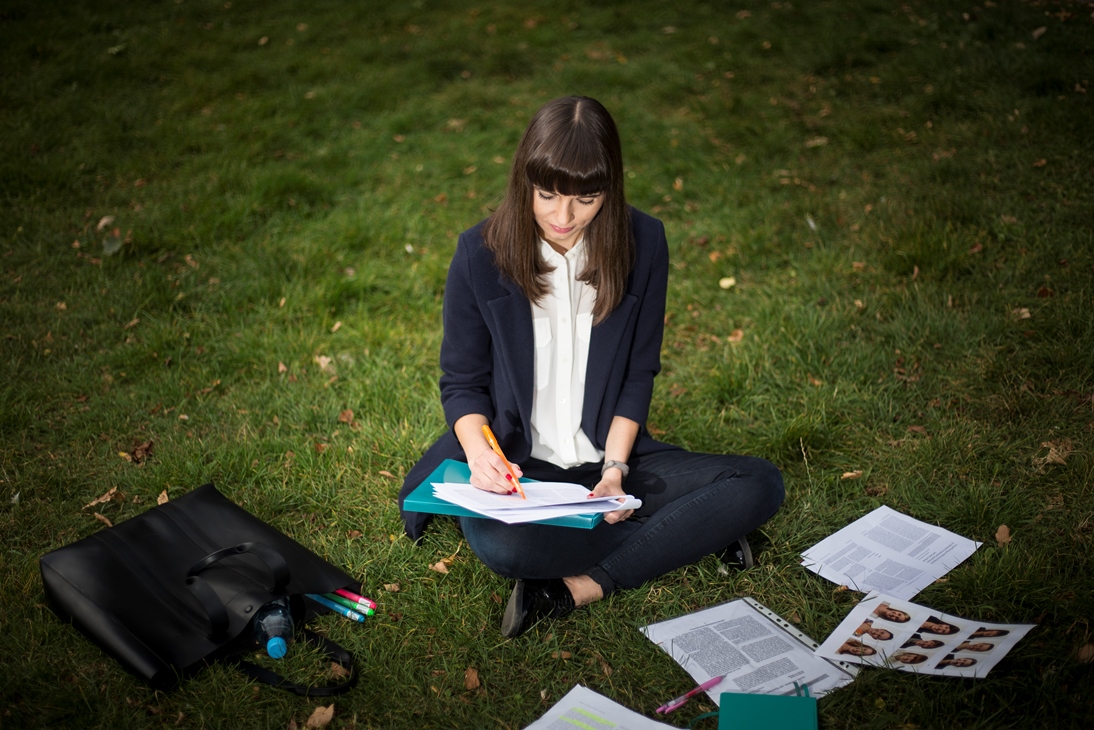 Monika Wróbel siedzi po turecku na trawie, z notesem na kolanach. W okół na trawniku leżą rozrzucone wydruki i torebka.