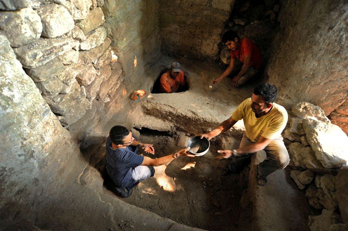 Zdjęcie przedstawia wnętrze kamiennego pomieszczenia. W podłożu znajdują się dwa płytkie wykopy - mniejszy i większy. Przy wykopach pracuje czterech mężczyzn.