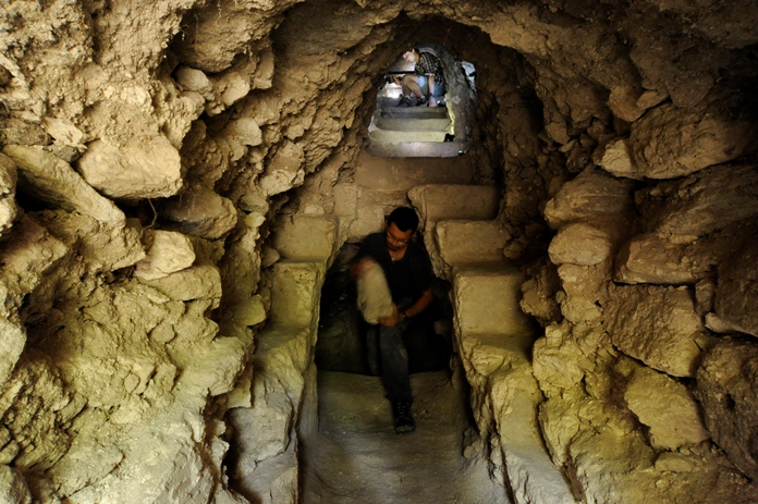 Wnętrze wąskiego, kamiennego pomieszczenia, przypominającego tunel. Tunelem idzie mężczyzna, trzymający w rękach przedmiot wykonany z kamienia. W tle, w innym pomieszczeniu, znajdują się dwie osoby, zaglądające do tunelu.
